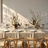 A styled dining table set for six with white plates, clear glassware, silver candle holders, and neutral beige table linens, accented by two large dried floral centerpieces. The table is surrounded by light wood chairs with woven seats against a warm off-white wall with soft sunlight and leaf shadows.