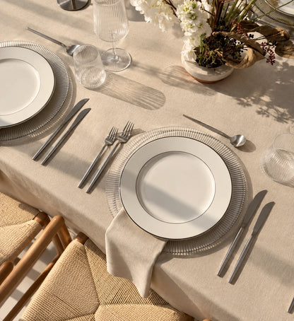 A styled dining table setting with white ceramic plates, silver cutlery, textured clear charger plates, a beige tablecloth, and a neutral woven chair. Natural sunlight casts soft shadows across the table.