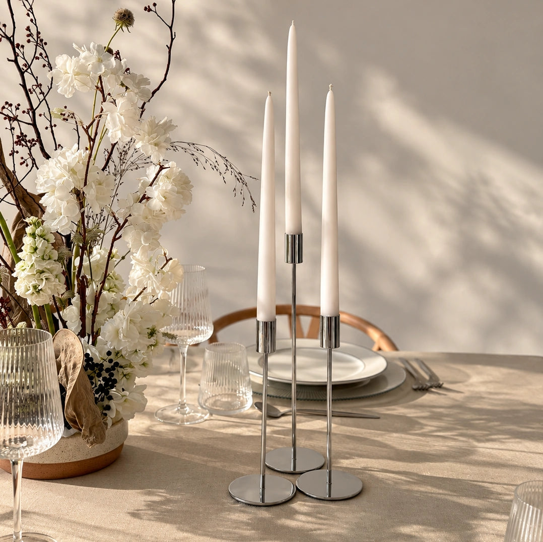 Three white taper candles in a slim silver metal candelabra set on a beige dining table, styled with a floral centerpiece, glassware, and plates in soft natural light.