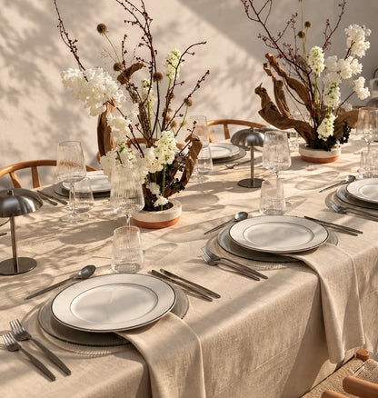 A styled dining table set with a beige linen tablecloth, white plates with gray rims, silver flatware, clear glassware, and rustic floral centerpieces with branches and white flowers in a warm indoor setting.