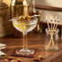 Cocktail with olives in a glass on a wooden bar with a cocktail recipe book and a jar of bar accessories.