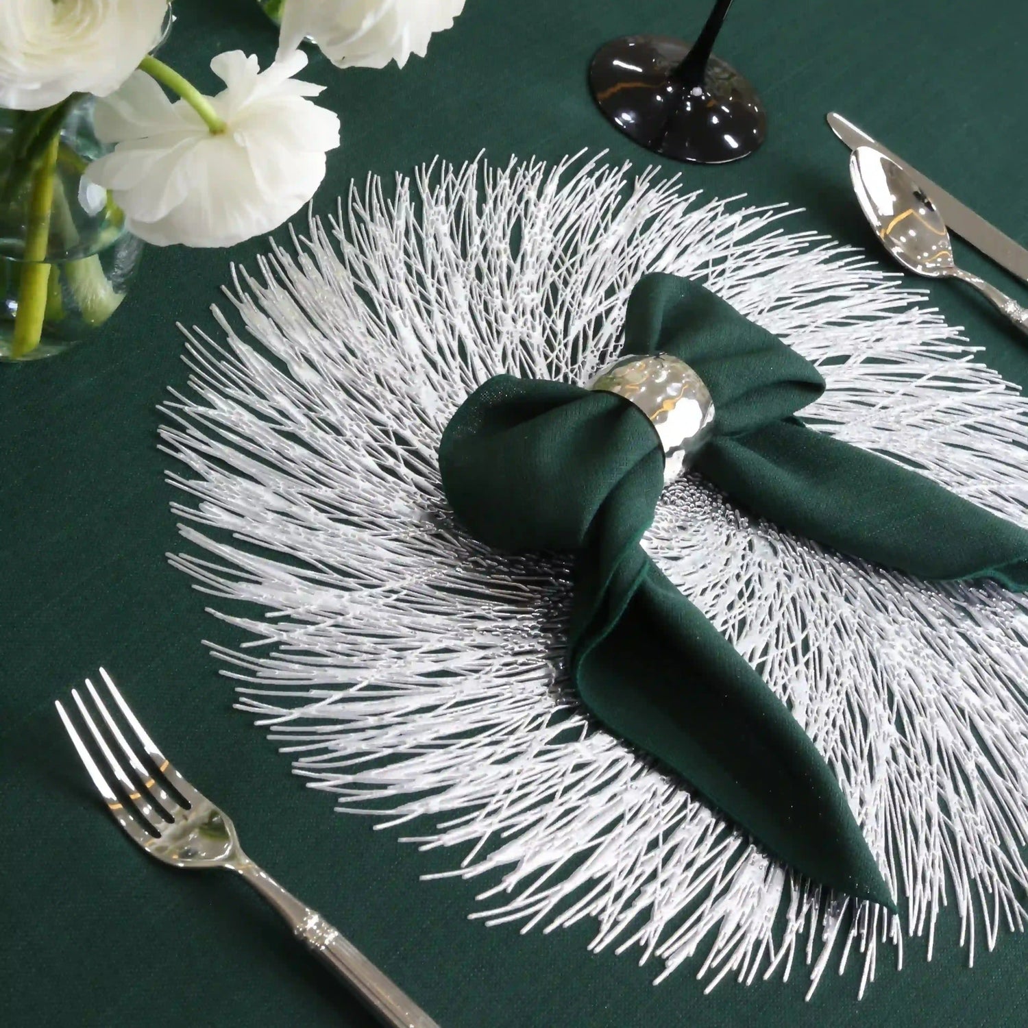 Silver Swirl Placemat on green table, featuring intricate spiral texture. Paired with a dark green napkin, silverware, and floral accents for elegant table setting.