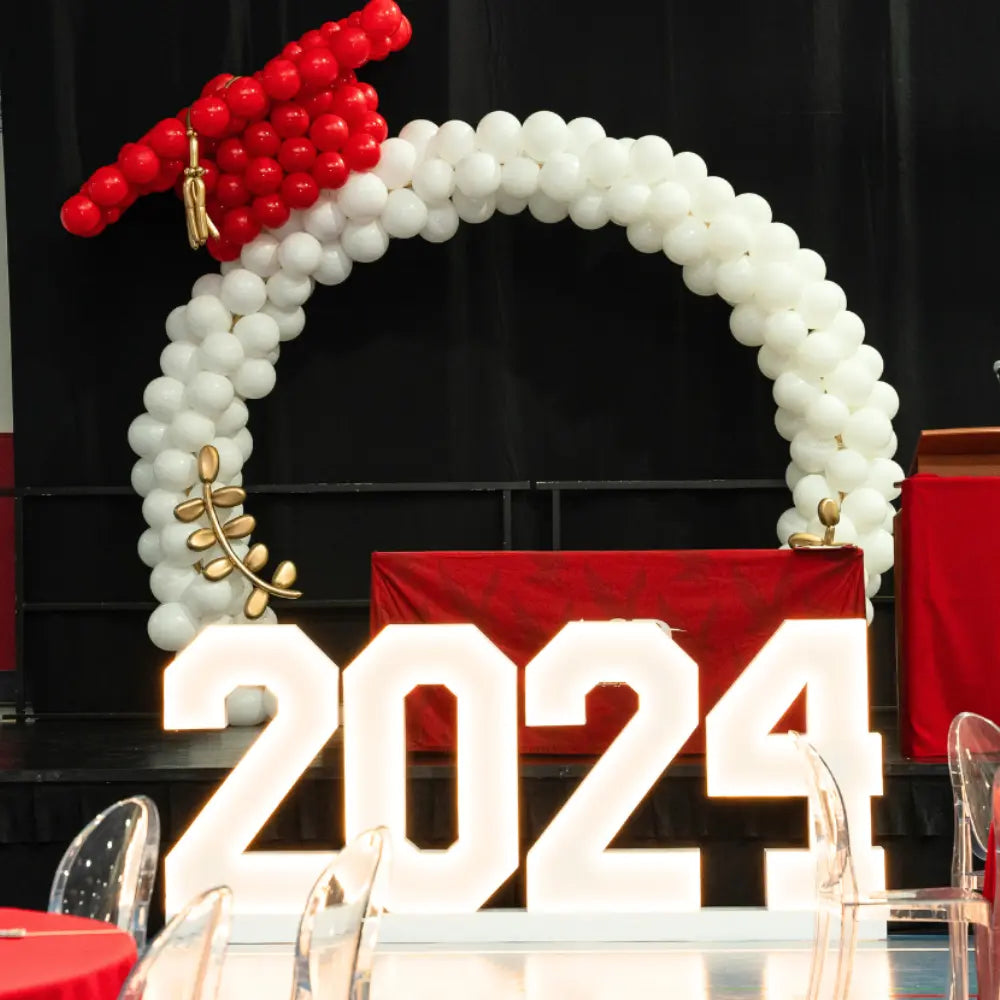 Graduation-themed arch with red cap, white balloons, and '2024' sign. Standing Letters and Numbers