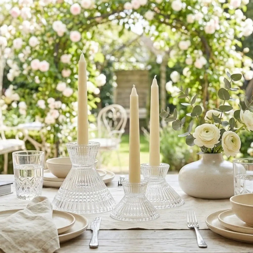 Elegant outdoor table setting with tall candles, candleholders, flowers, and cutlery in a garden.