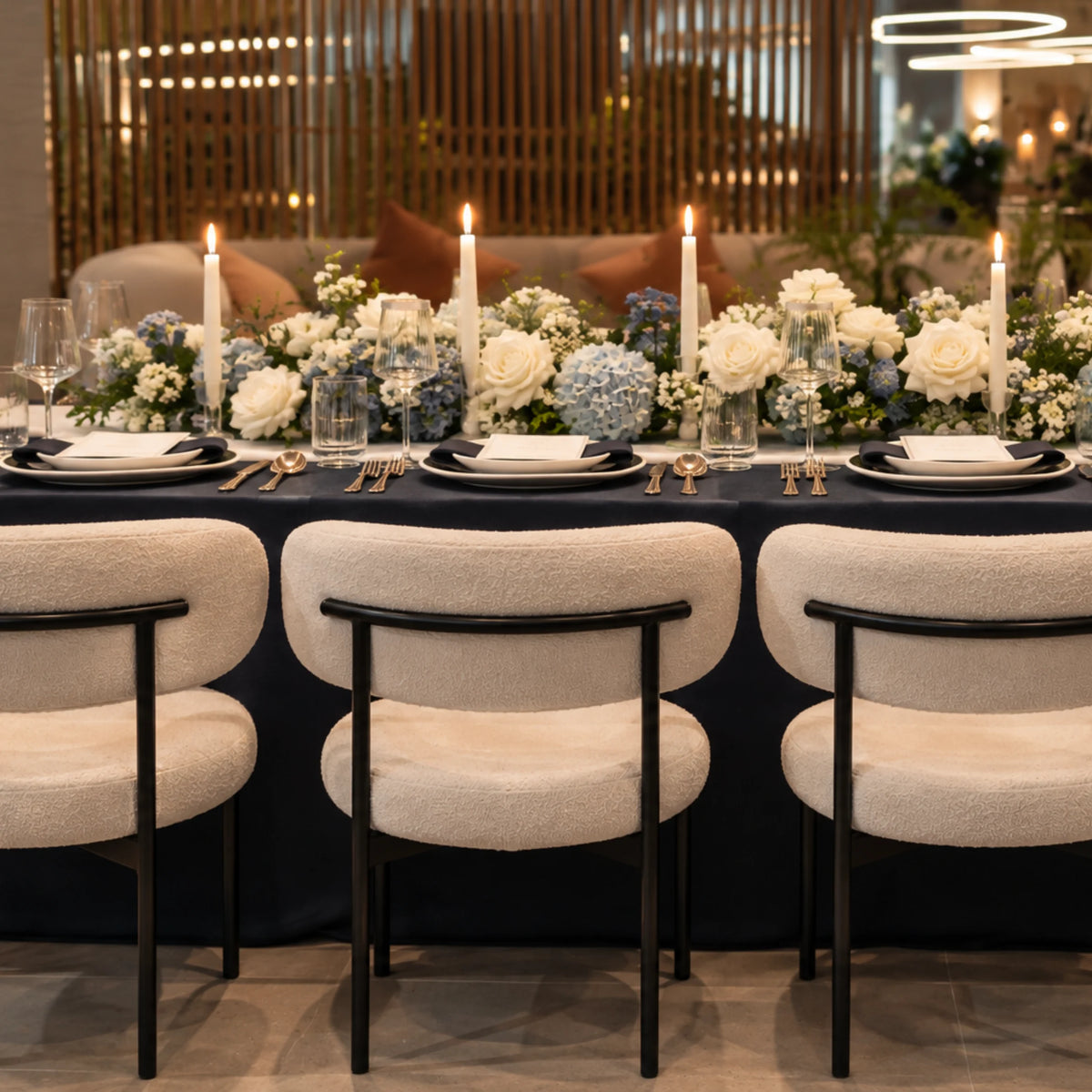 A styled dining table setup in a modern interior, featuring white bouquets with blue hydrangeas, lit taper candles, black plates, and three upholstered dining chairs with cream fabric and black frames in the foreground.