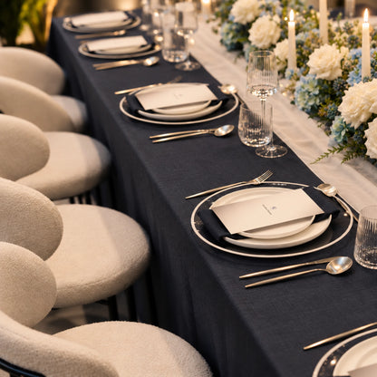 A long dining table set for guests with a dark navy tablecloth, white plates, silver cutlery, clear drinking glasses, and a floral centerpiece with lit candles. White upholstered chairs line the table.