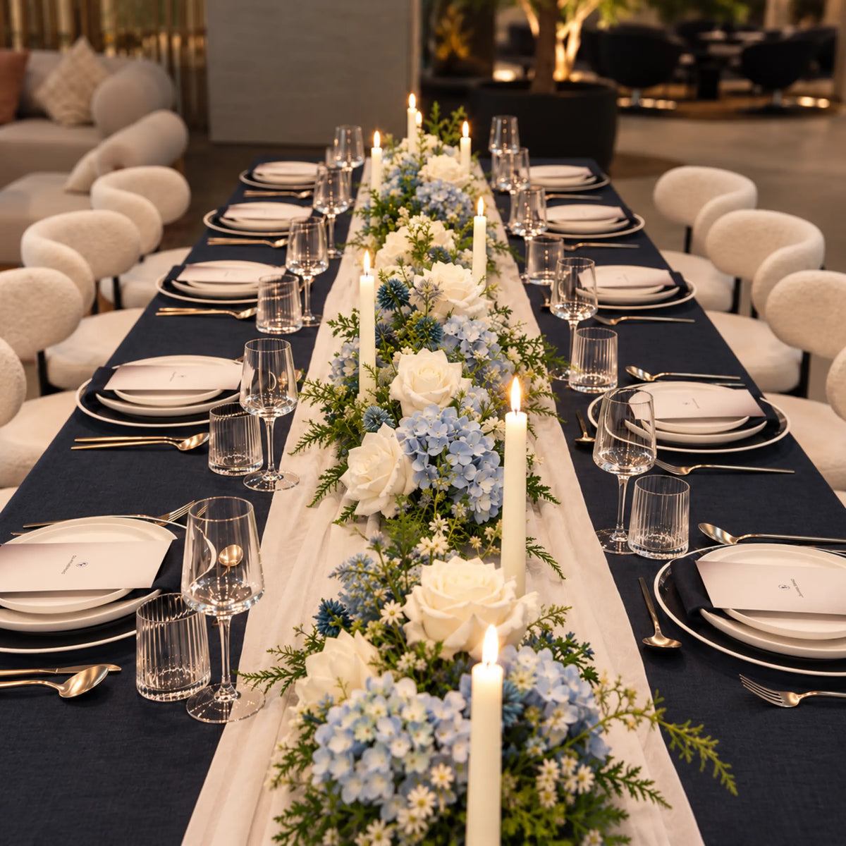 A long banquet table set for guests with navy and white tablecloths, white plates, clear glassware, and a floral centerpiece of white and light blue roses with green foliage and lit taper candles.