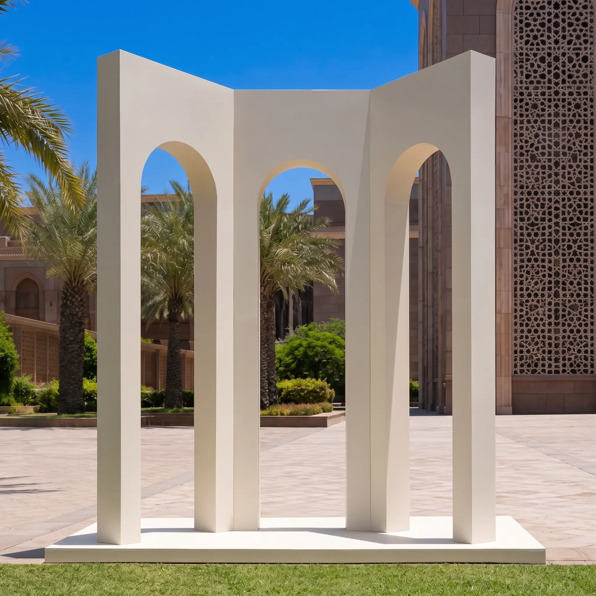 A modernist white concrete sculpture featuring three tall arches on a square base, set outdoors on a lawn with a paved courtyard, palm trees, and a building facade in the background.