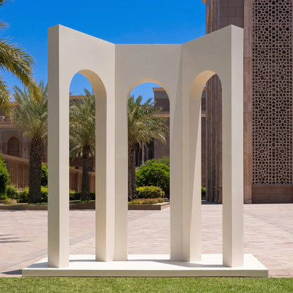 A modernist white concrete sculpture featuring three tall arches on a square base, set outdoors on a lawn with a paved courtyard, palm trees, and a building facade in the background.