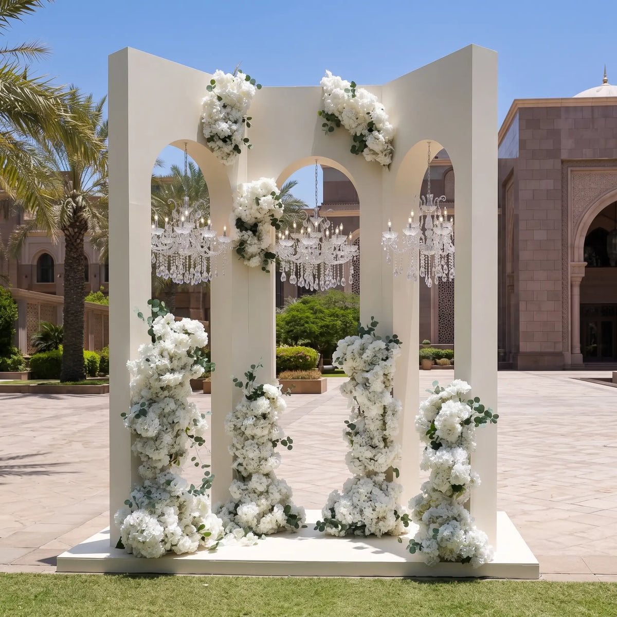 A large white arched floral backdrop stand decorated with cascading white flowers and greenery, with hanging crystal chandeliers visible behind it in an outdoor courtyard setting.