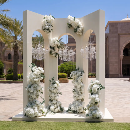 A large white arched floral backdrop stand decorated with cascading white flowers and greenery, with hanging crystal chandeliers visible behind it in an outdoor courtyard setting.