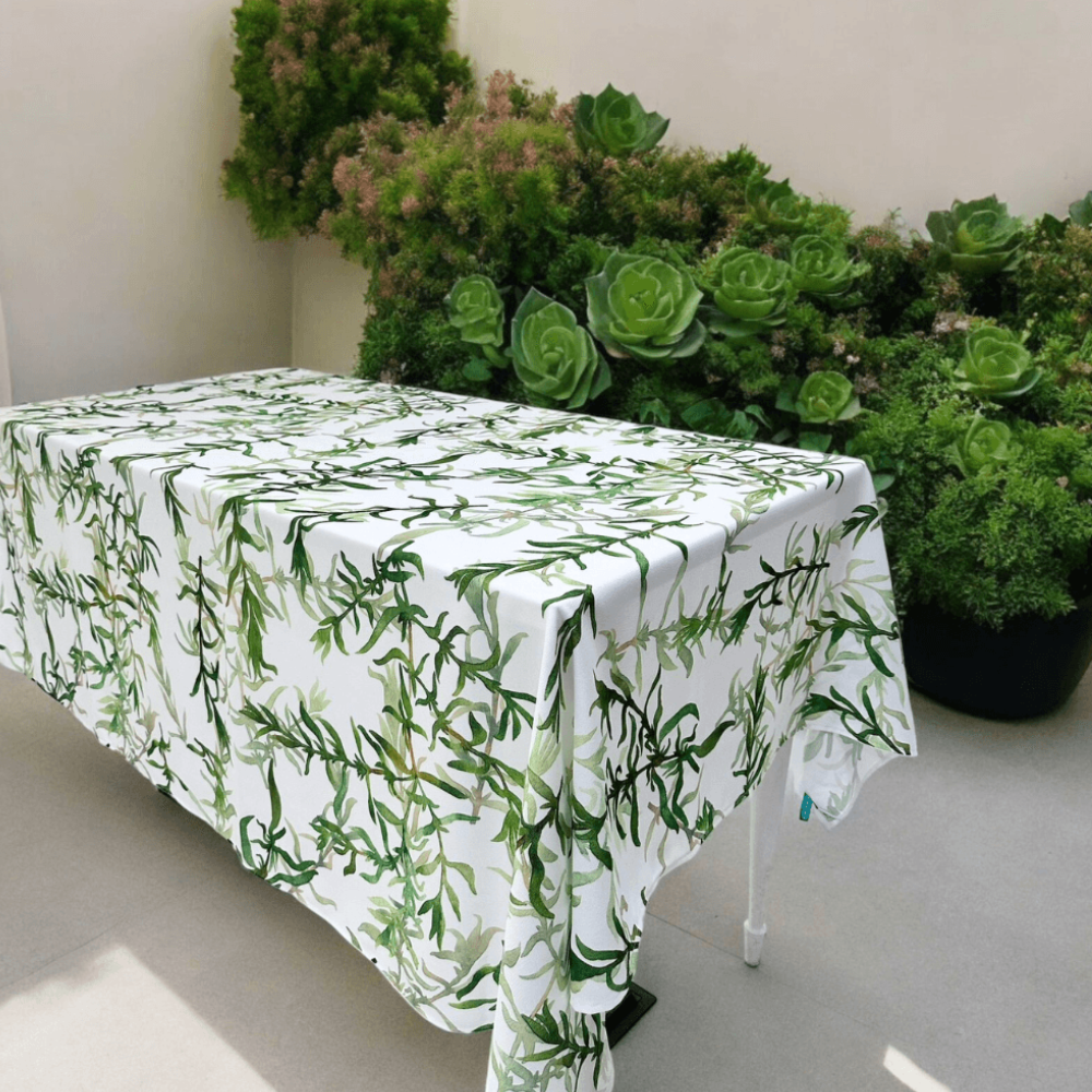 Gardenia Linen Tablecloth (1pc) on a table, showcasing a white polyester fabric with green leaf prints, complemented by houseplants in the background.