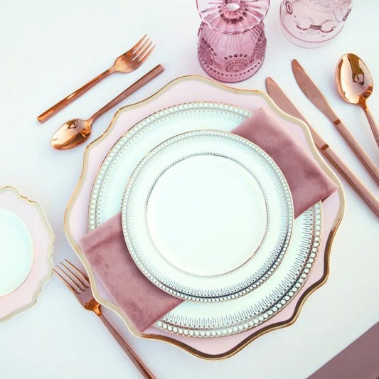 Pink Lotus Collection: Elegant table setting with scalloped charger plates, mauve-pink napkin, rose-gold flatware, and pink glasses on a white tablecloth.