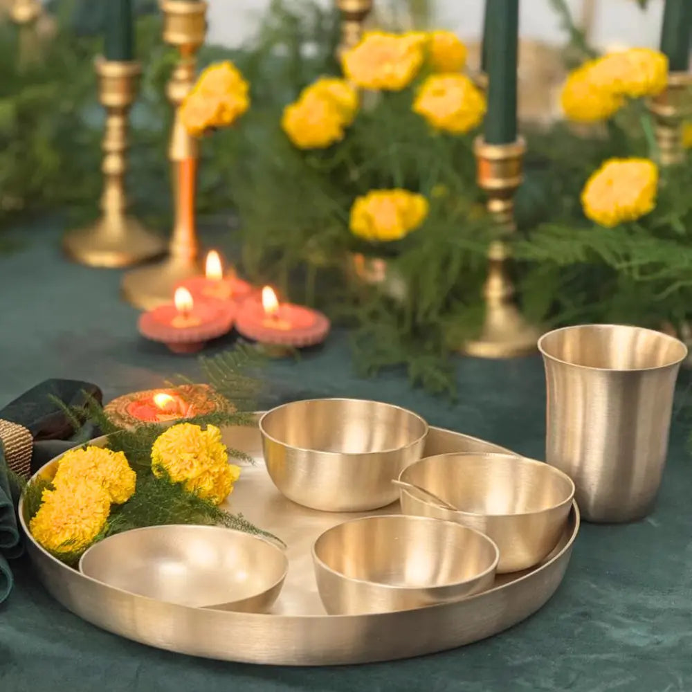 A brass-colored metal ritual tray set with five small bowls and a tall cup, styled on a dark green tablecloth with yellow flowers, candles, and green candlesticks in the background.