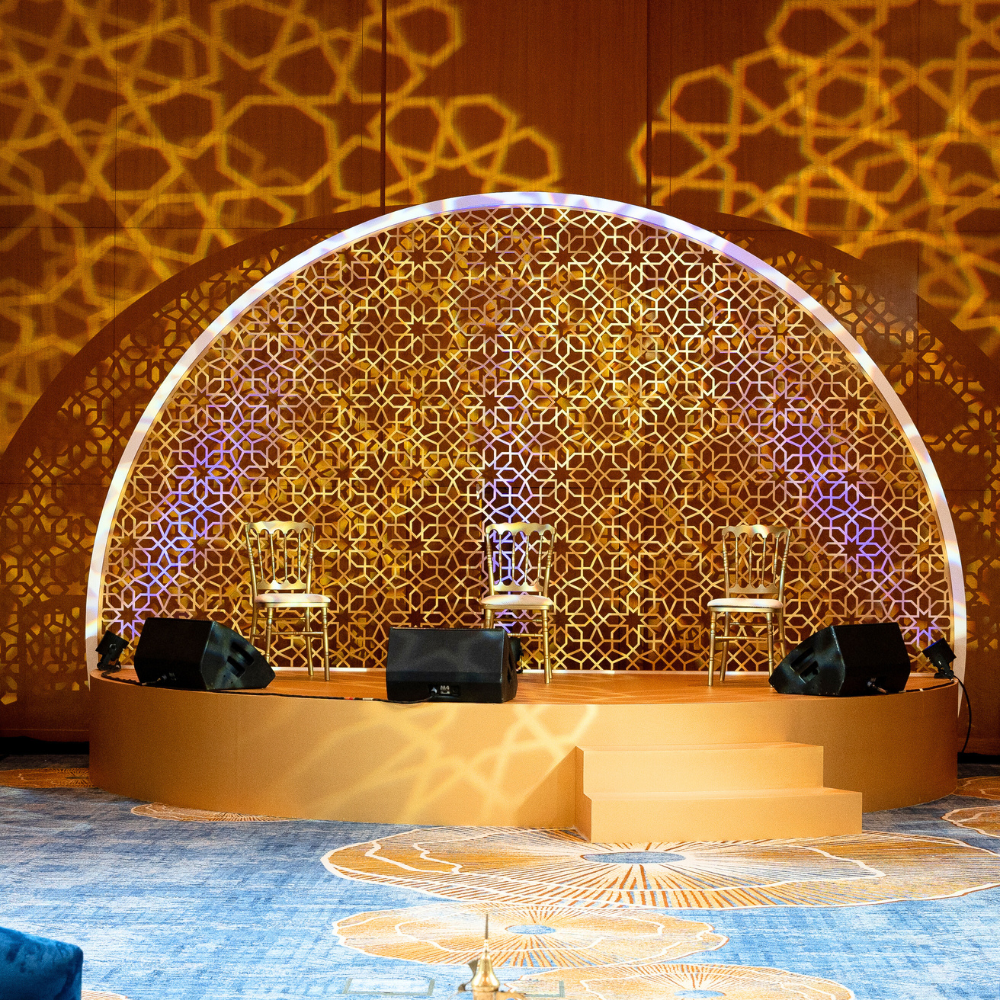 Arabesque Stage with gold backdrop, designed for themed events and weddings, showcasing elegant indoor setup with chairs and lighting, available at Party Social.