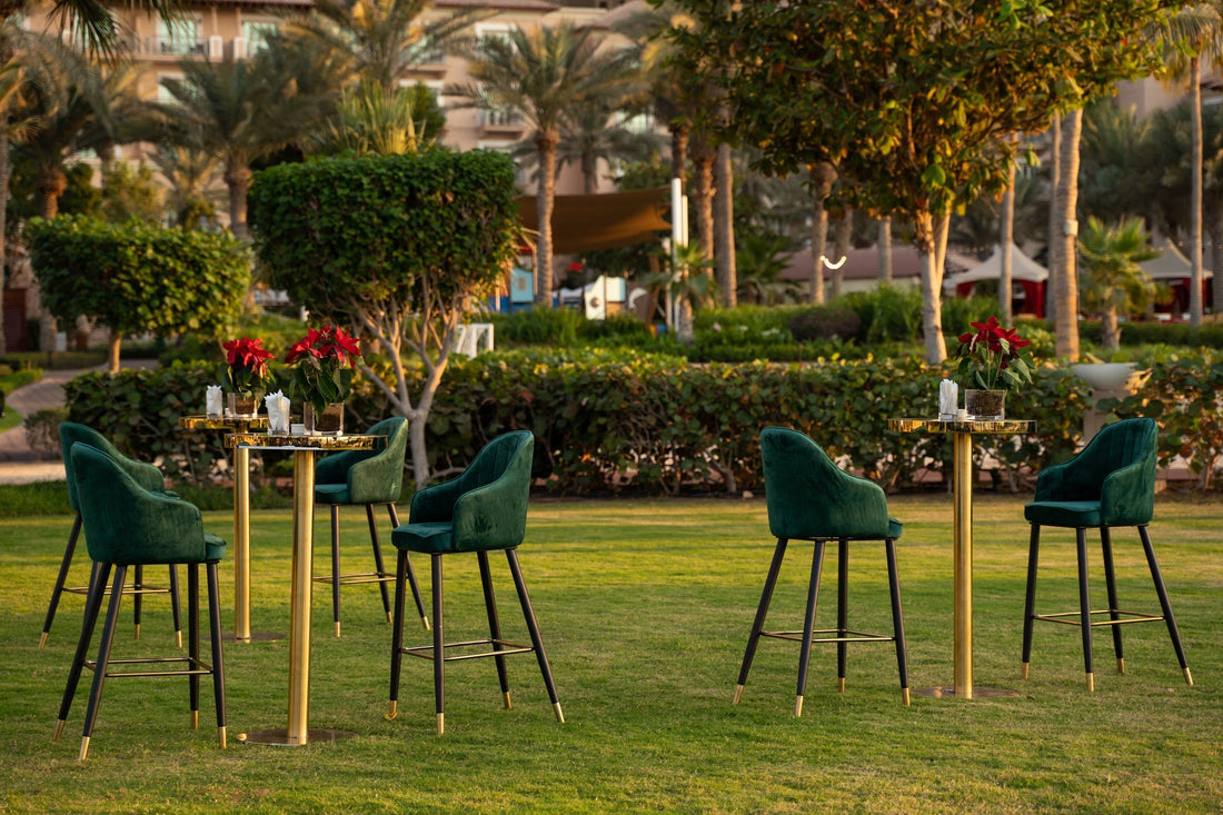 VELVET STOOL ROYAL GREEN, featuring velvet fabric and black and gold legs, ideal for weddings and events, set on a grassy field with outdoor furniture.