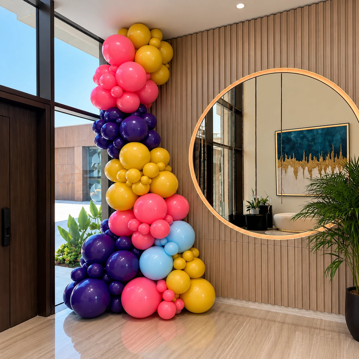 A decorative cluster of inflated balloons in pink, yellow, purple, and light blue arranged in a curved column inside a modern interior with wood slat walls, a large round mirror, and a potted plant.