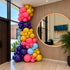 A decorative cluster of inflated balloons in pink, yellow, purple, and light blue arranged in a curved column inside a modern interior with wood slat walls, a large round mirror, and a potted plant.
