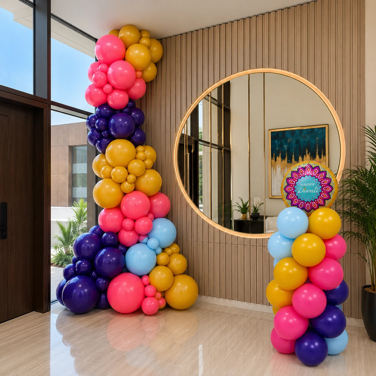 A colorful balloon garland decoration set in a modern indoor space, featuring a tall column of balloons in pink, yellow, purple, and light blue next to a smaller matching column with a decorative sign that reads &