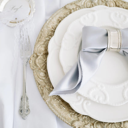 White Embossed Collection plates stacked elegantly on a gold charger, with a silvery-blue napkin and patterned silverware, ideal for sophisticated event settings.