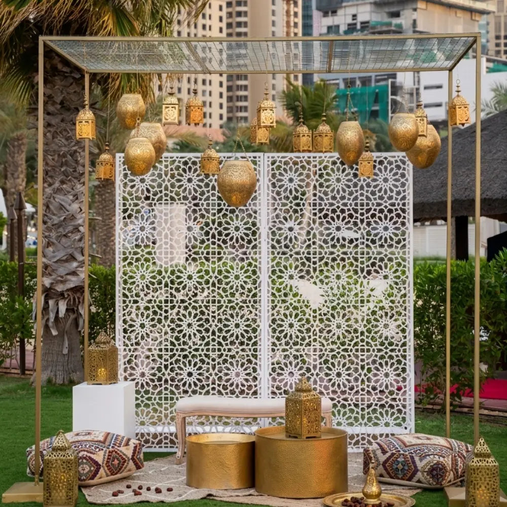 WHITE ARABESQUE backdrop with geometric floral patterns, complemented by an elegant outdoor lounge setup featuring gold tables, lanterns, and plush poufs for event decor.