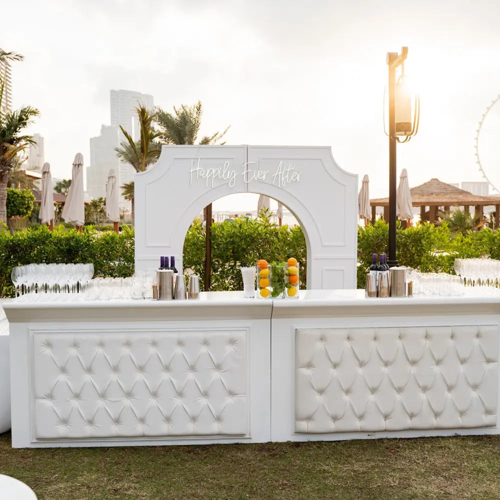 WHITE LEATHER BAR UNIT with tufted front, set for an outdoor event with glasses, shakers, and vases, against a lush garden backdrop and decorative arch.