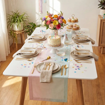 WHITE RECTANGULAR TABLE set for a festive party with a pastel table runner, cake, cupcakes, flower bouquet, gold flatware, and champagne glasses.