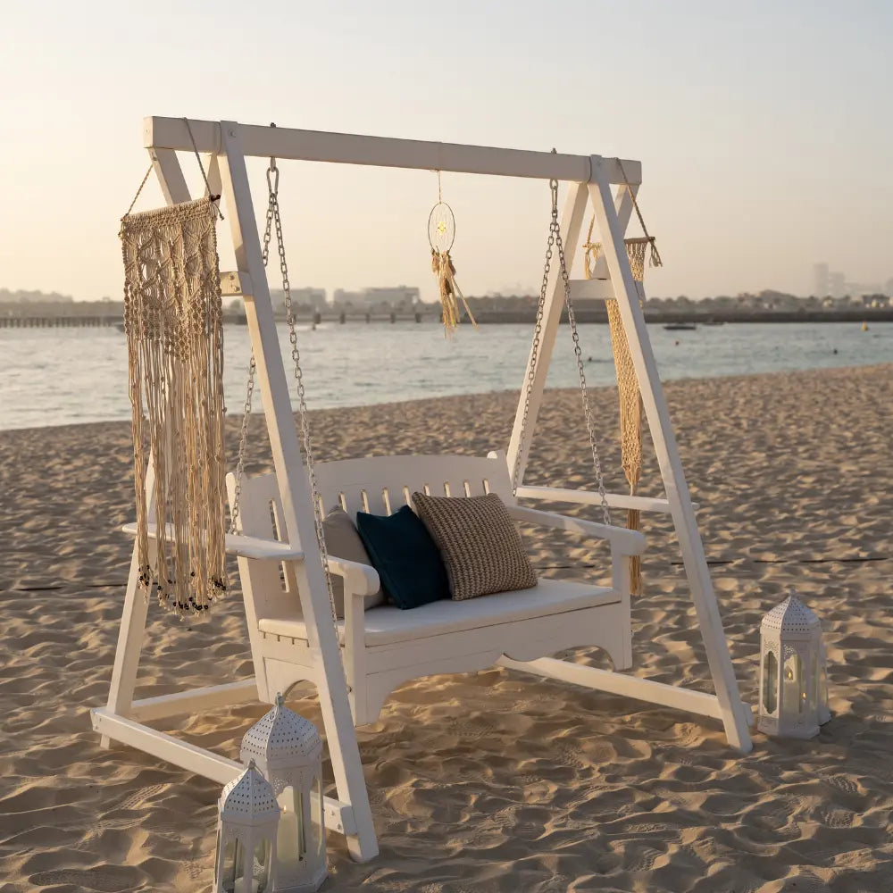 WHITE WOODEN SWING on sandy beach with boho decor, including macramé and dreamcatcher, perfect for event props. Two lanterns accentuate the setup.