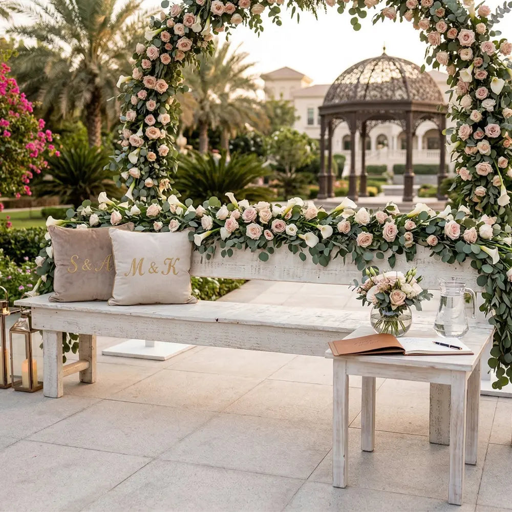 White wood bench with eucalyptus and rose garland, featuring monogrammed pillows. Complements rustic event settings, ideal for wedding welcome or photo spots.