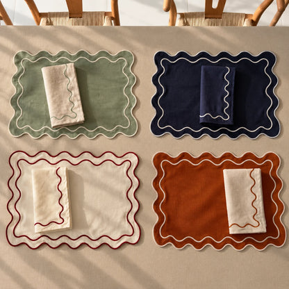 Four scalloped fabric placemats arranged on a beige table, each paired with a matching folded napkin. The placemats come in sage green, navy blue, cream with red trim, and rust orange with white trim.