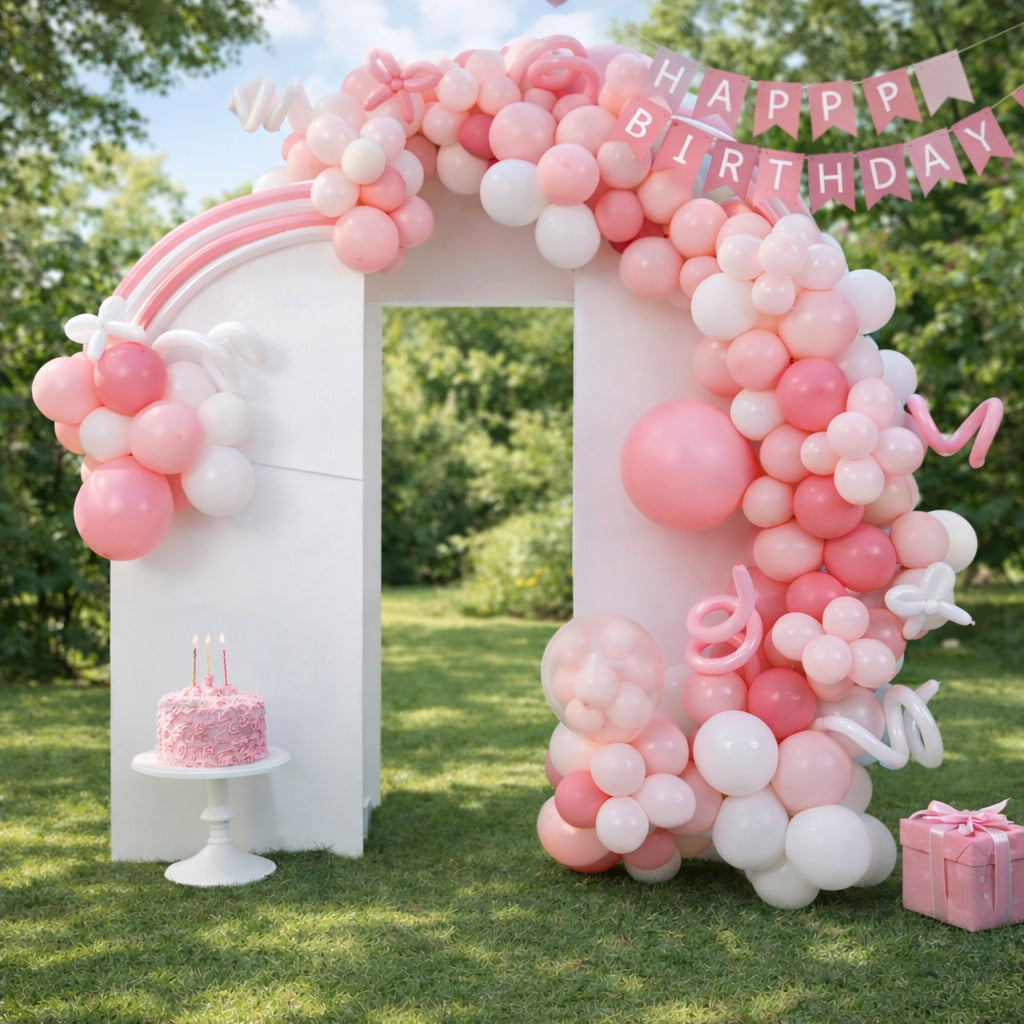 Balloon arch with pink and white balloons, 'Happy Birthday' banner, cake, and gift on grass