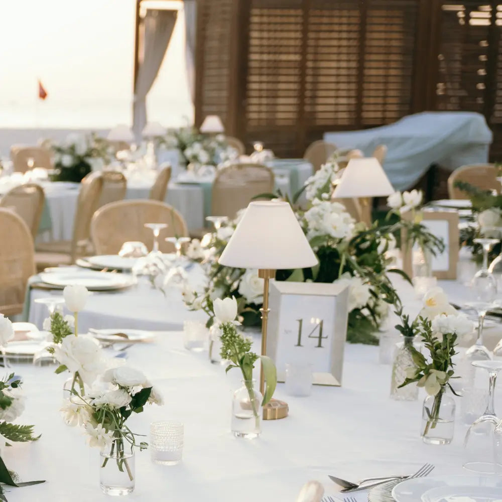 Elegant outdoor wedding reception with tables set for guests, featuring white tablecloths, floral arrangements, and decorative lamps.