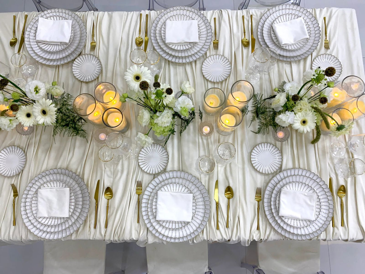 Classic Cream-Dining Setup: Elegantly arranged table for eight, featuring scalloped plates, gold cutlery, glassware, ghost chairs, floral centerpieces, and lit candle vases.