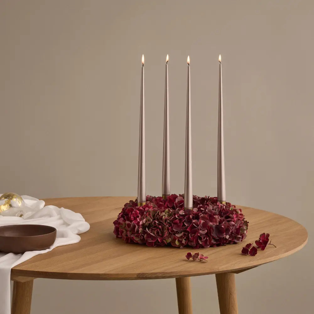 A floral candle holder centerpiece on a round wooden table, featuring five tall taper candles standing upright in a ring of dark red and burgundy dried flowers, with a neutral beige background.