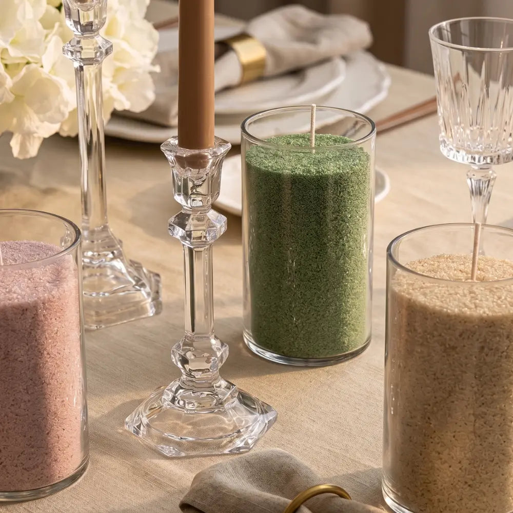 Clear glass candle holders and tumbler glasses containing textured wax candles in green, pink, and beige tones, styled on a beige tablecloth with a candlestick, white flowers, and folded napkins.