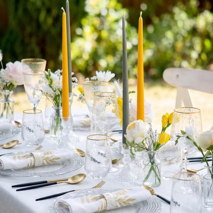 A styled outdoor dining table setting with white tablecloths, clear textured glassware, gold-toned cutlery, folded white napkins with gold leaf patterns, and yellow and gray taper candles in a softly blurred garden background.