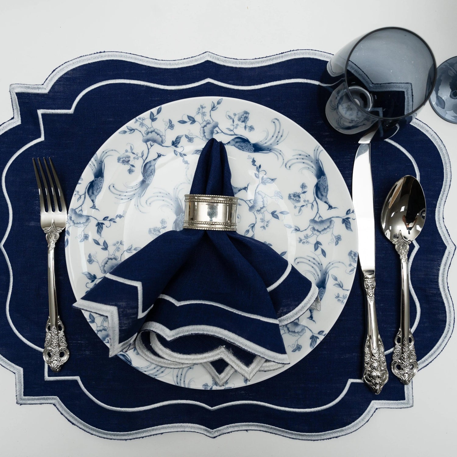 Top-down view of Navy Scalloped Linen Placemats in an elegant table setting with matching napkin, silver cutlery, and a smoky glass goblet.