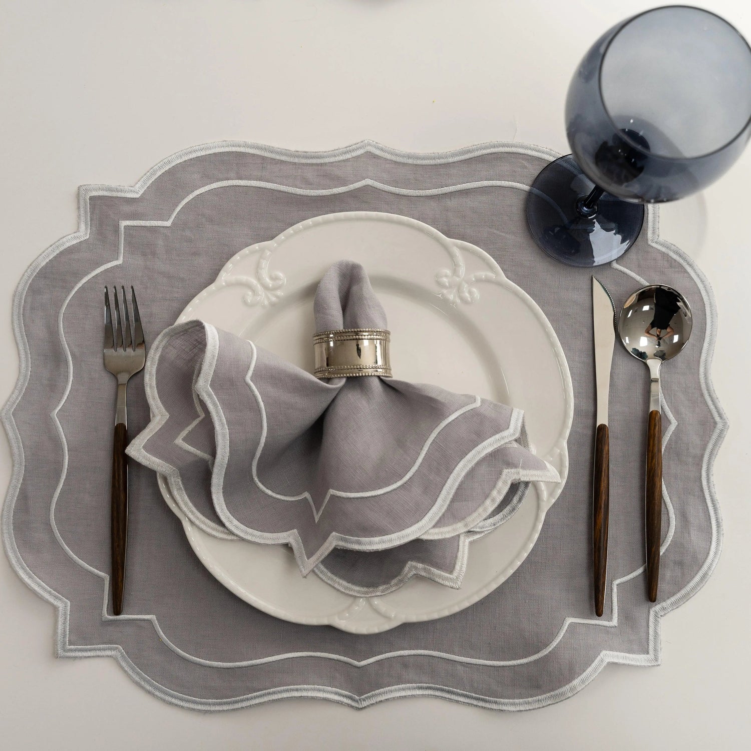 Light Grey Scalloped Linen Placemats - Set of 4 on a white tabletop with matching napkin, silver ring, white plate, dark-wood flatware, and smoke-blue wine glass.