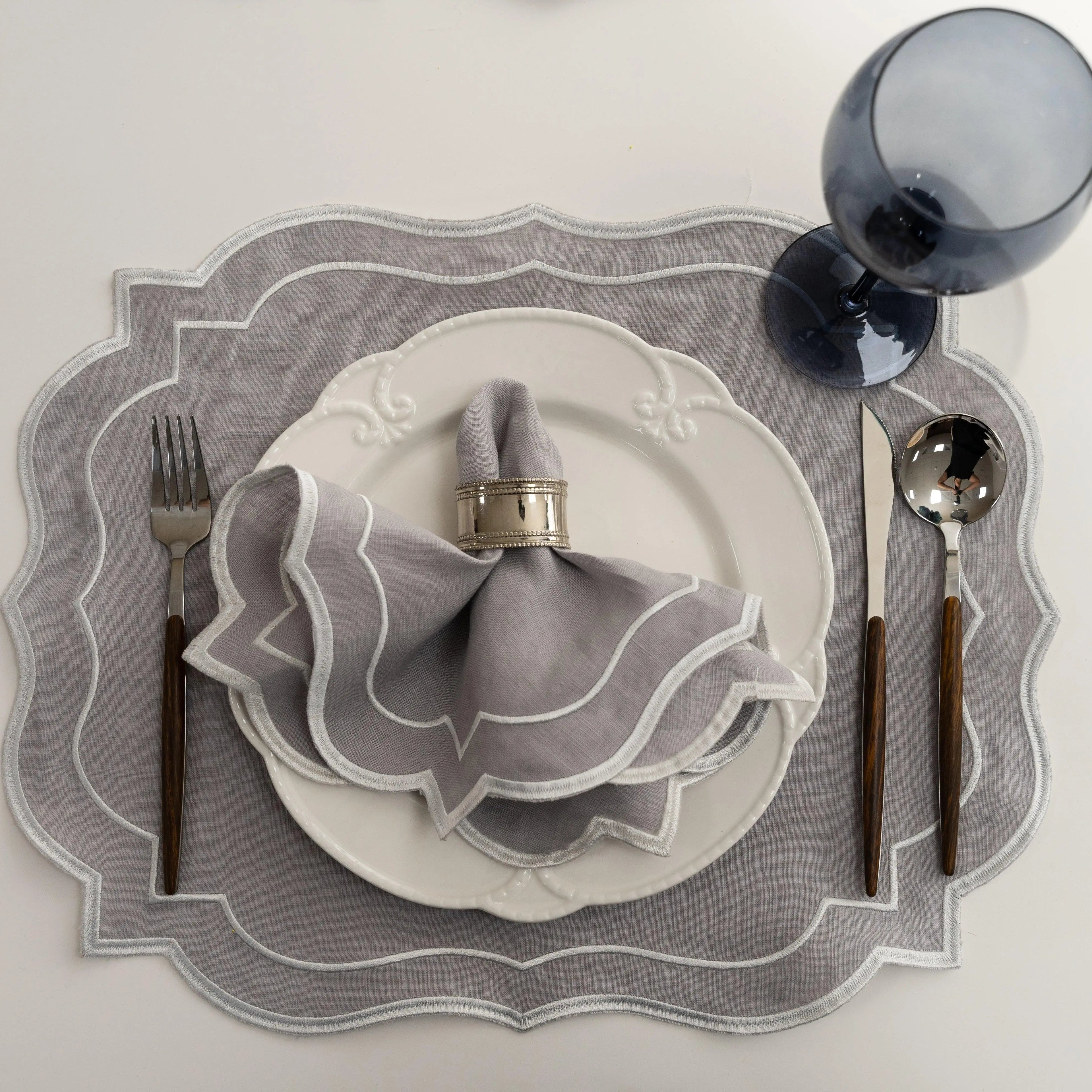 Light Grey Scalloped Linen Placemats - Set of 4 on a white tabletop with matching napkin, silver ring, white plate, dark-wood flatware, and smoke-blue wine glass.